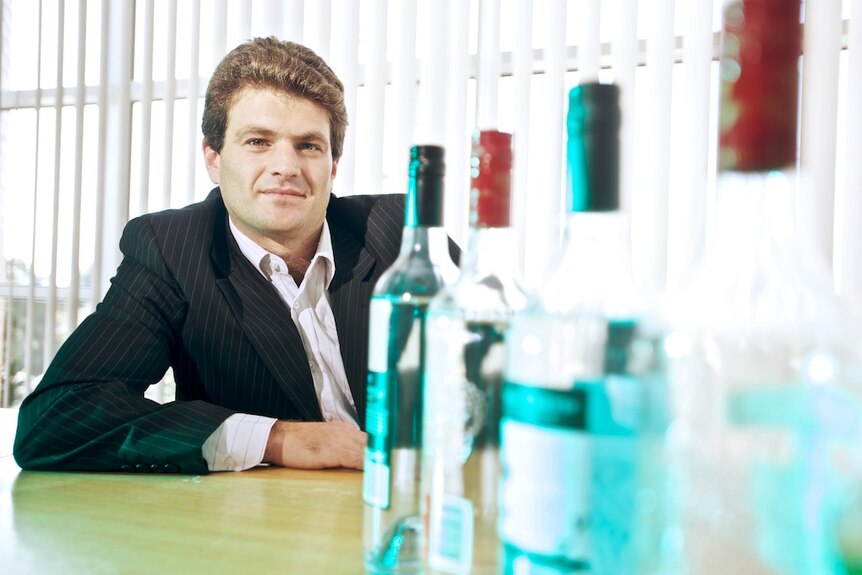 Empty alcohol bottles in front of a man at a desk. He has curly auburn hair and is wearing a dark suit and white shirt, no tie