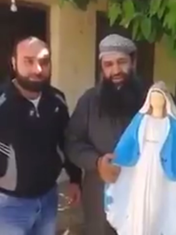 Still from video of a Syrian man breaking a blue and white Virgin Mary statue. He has a beard and stands beside another man.