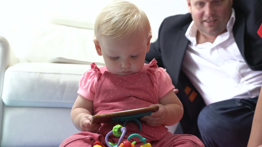 One-year-old Stella sitting on the floor looking at a mobile phone, while her father sits behind her