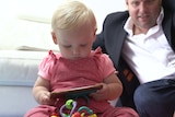 One-year-old Stella sitting on the floor looking at a mobile phone, while her father sits behind her