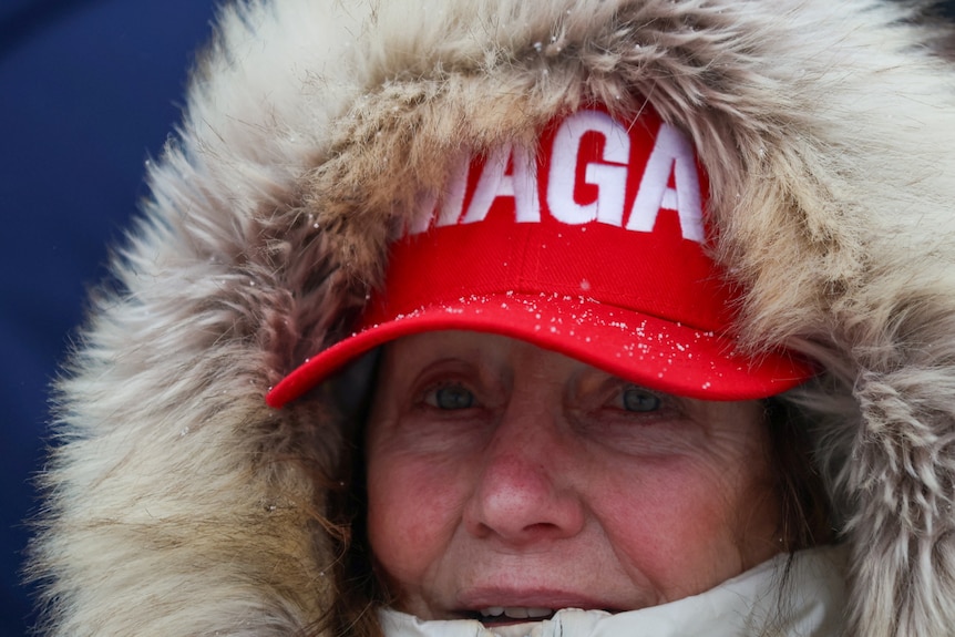 A woman wearing a fur coat and a MAGA hat waits outside.