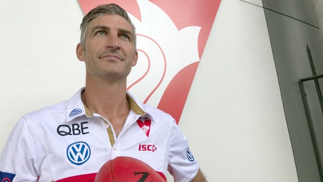 Brett Kirk holds a football while standing in front of a Sydney Swans logo.