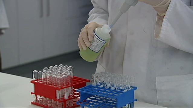 A hand uses pipette in bottle in front of rack of test tubes in a science laboratory