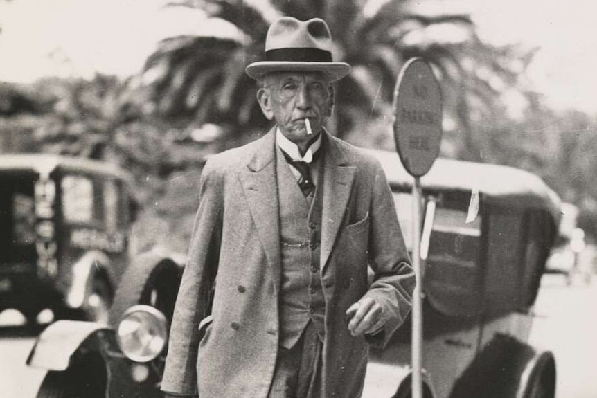 Portrait photo of a man in a suit and a fancy hat walking along the sidewalk with a cigar in his mouth.