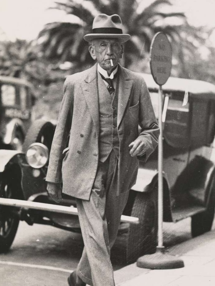 Portrait photo of a man in a suit and a fancy hat walking along the sidewalk with a cigar in his mouth.