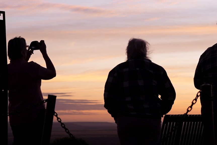 a group of people stand at a mountaintop lookout at sunrise taking photos