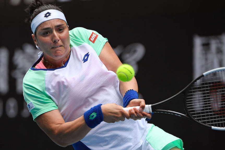 A tennis player grimaces as the ball comes off her racquet when she hits a backhand.