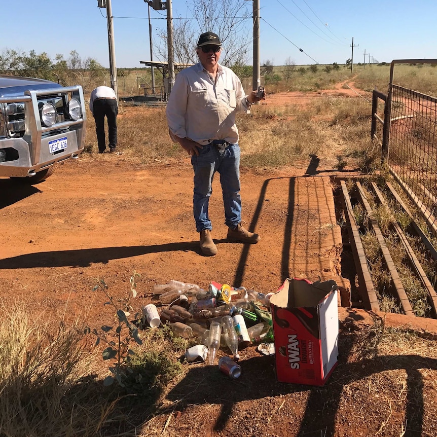 A man stands holding a broken lock behind piles of rubbish and empty beer cartons on his station