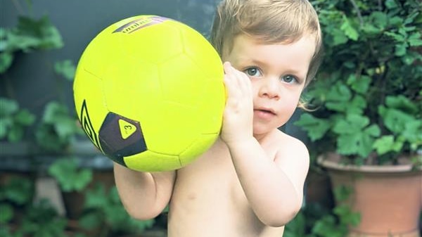 A toddler holds a bright yellow ball while wearing a cloth nappy in the backyard.