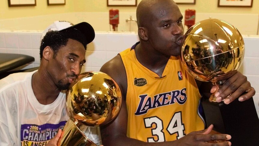 Kobe Bryant in a white shirt and cap next to Shaquille O'Neal in a Lakers jersey kissing NBA Championship trophies
