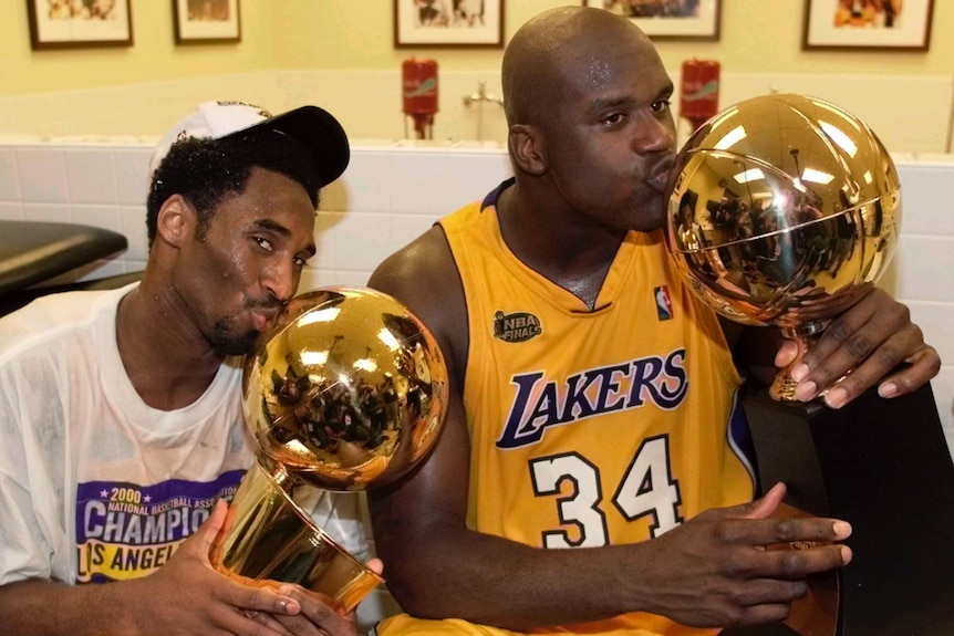 Kobe Bryant in a white shirt and cap next to Shaquille O'Neal in a Lakers jersey kissing NBA Championship trophies