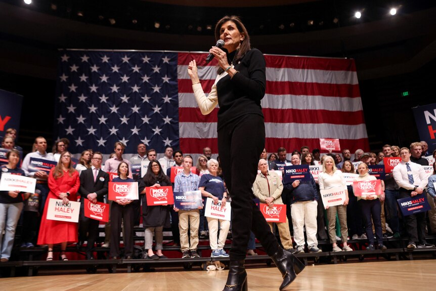 A middle-aged woman in a black and white outfits speaks into a microphone in front of supporters and a large American flag.