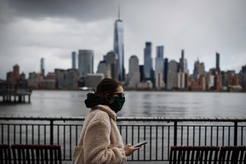 A woman in a coat and face mask walks past a river with the New York City skyline blurred in the background. She's holding phone