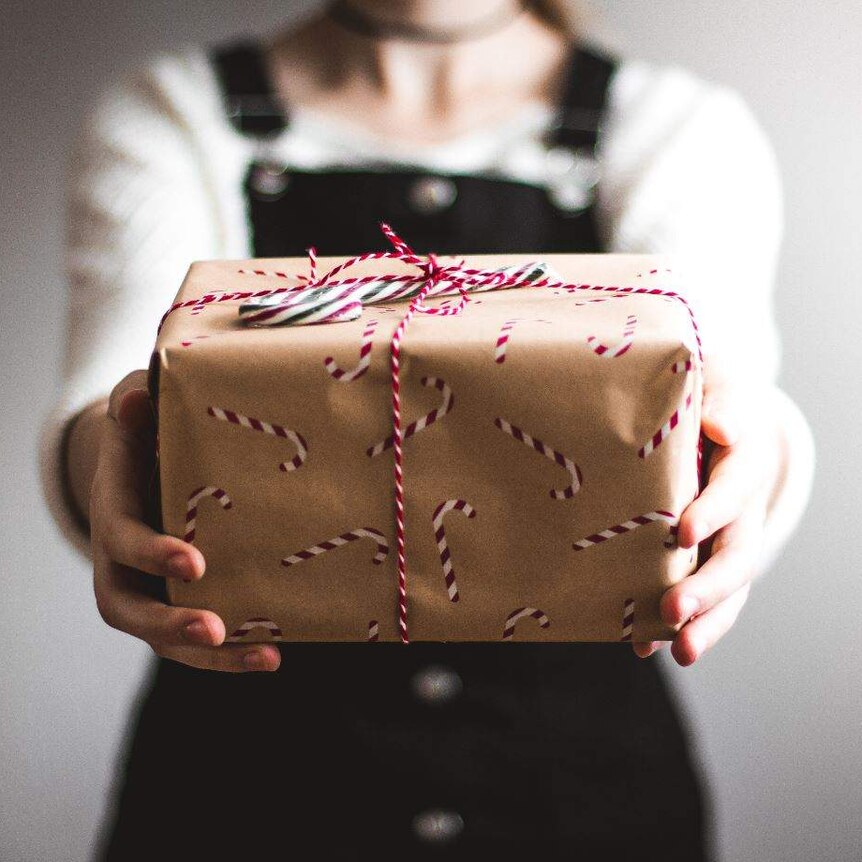 A person holding out a box wrapped in Christmas paper with a red bow and a candy cane on top.