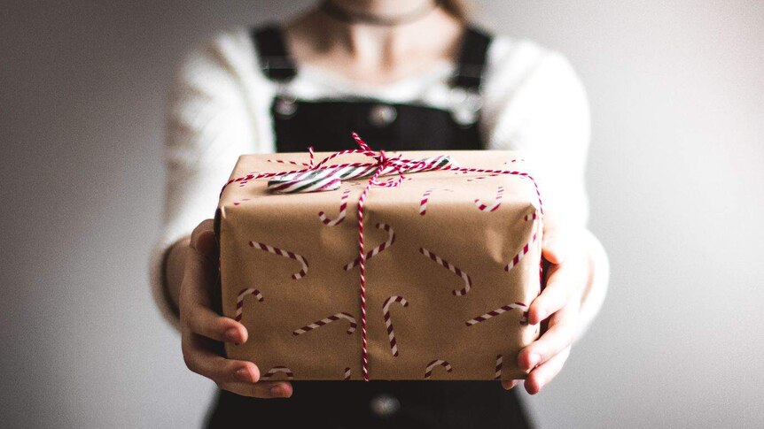 A person holding out a box wrapped in Christmas paper with a red bow and a candy cane on top.