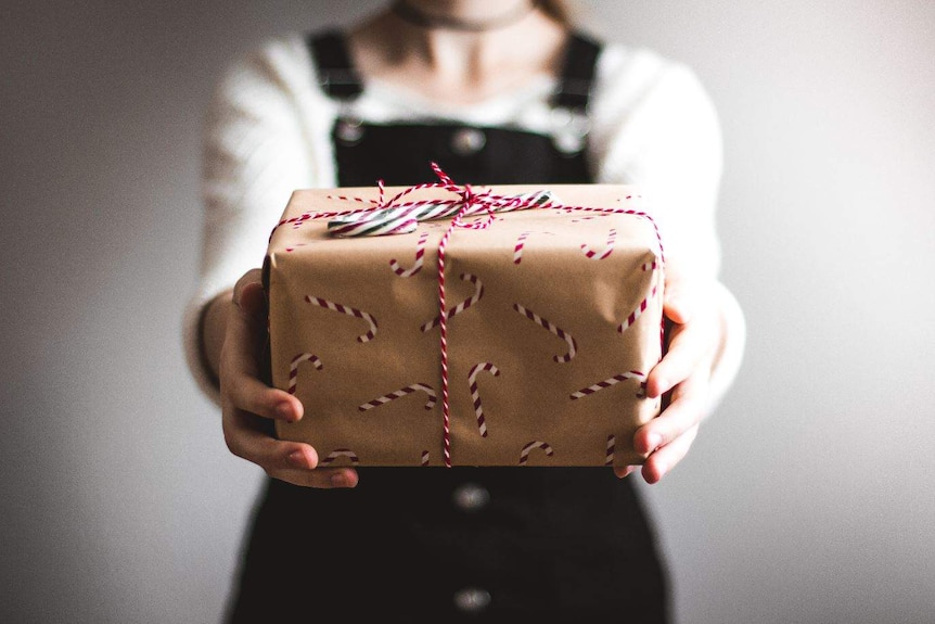 A person holding out a box wrapped in Christmas paper with a red bow and a candy cane on top.