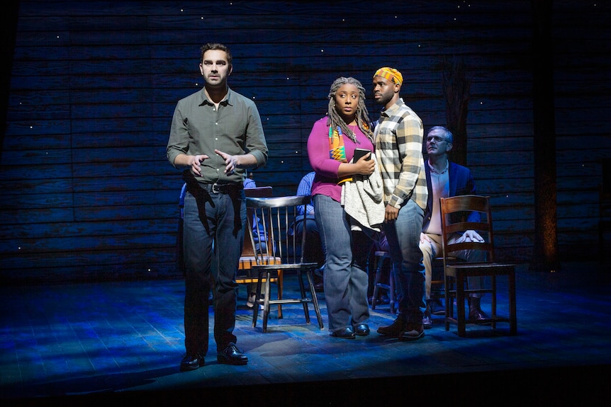 A man sings on stage while a man and a woman stands behind him