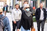 Woman with blue surgical mask walking down outdoor shopping mall, surrounded by several other people not wearing masks.