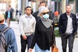 Woman with blue surgical mask walking down outdoor shopping mall, surrounded by several other people not wearing masks.