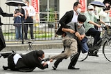 Two men grab a man running in a grey shirt and brown pants on a city street while bystanders watch on.