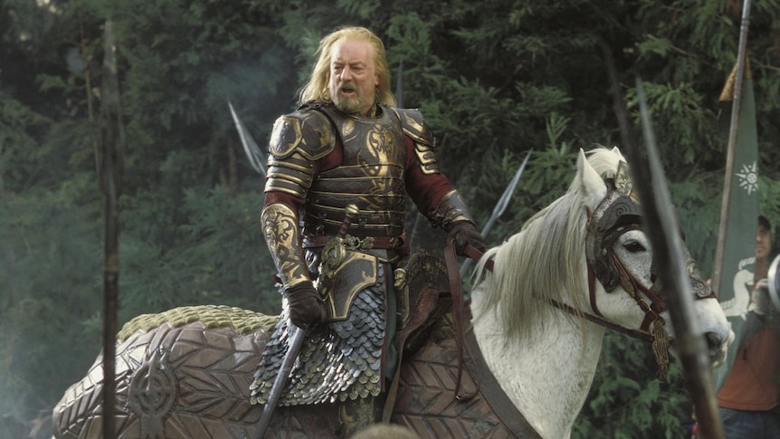 A blond-haired older man in impressive medieval armour rides a white horse through between soldiers with spears.