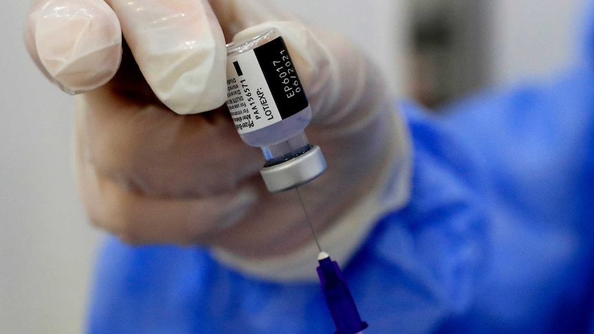 A gloved hand holds a Pfizer vaccine vial while inserting a syringe needle inside