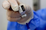 A gloved hand holds a Pfizer vaccine vial while inserting a syringe needle inside