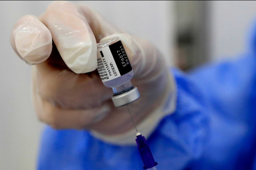 A gloved hand holds a Pfizer vaccine vial while inserting a syringe needle inside