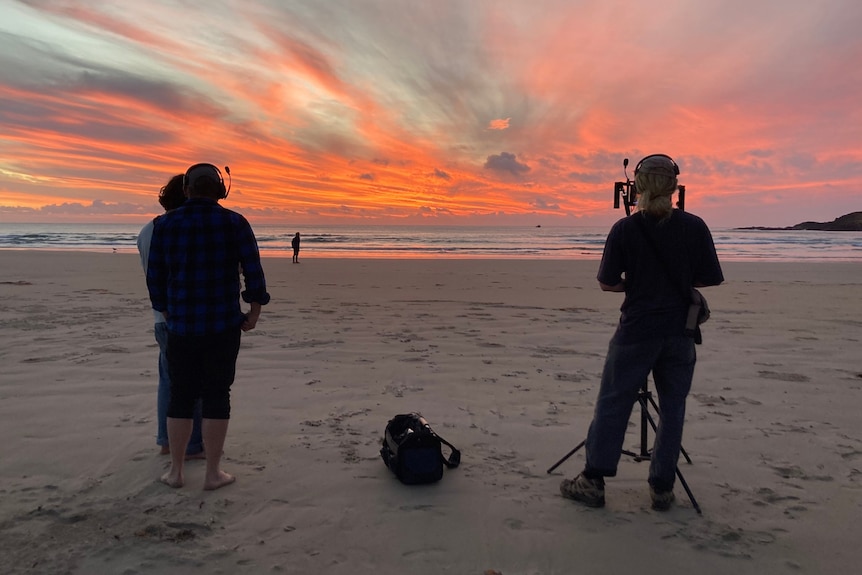 A cameraman stands on a beach with a red sky at sunset above 