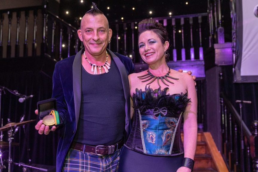 A smiling man and woman in striking attire, both with interesting hairdos, stand in front of a staircase next to a drumkit.