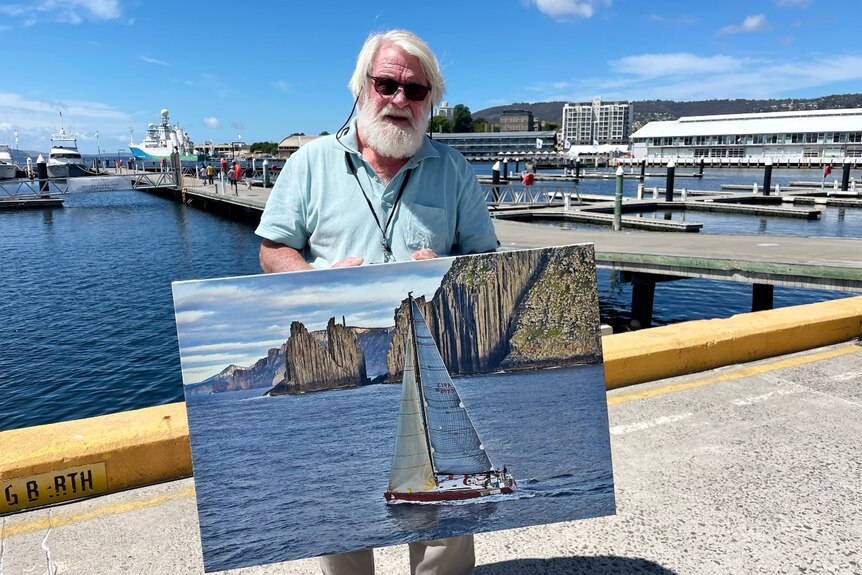 A man with silver hair and a beard holds a photo of a yacht sailing past cliffs