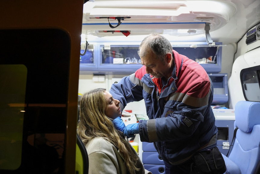 A young woman with long blonde hair sits in an ambulance as an older male paramedic treats her.