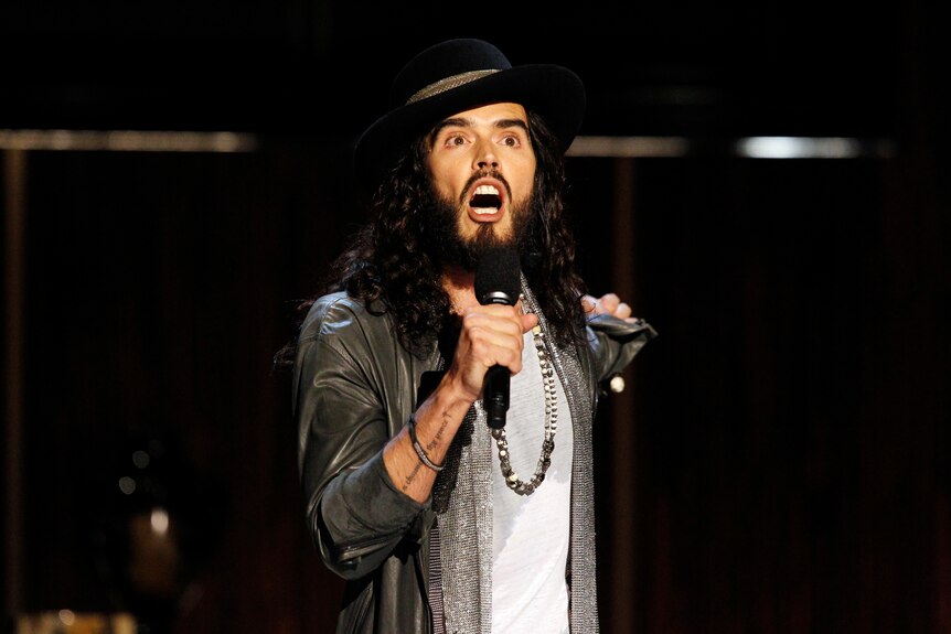 Russell Brand is pictured singing with a microphone in his hand.