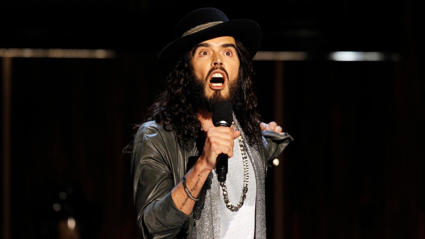 Russell Brand is pictured singing with a microphone in his hand.
