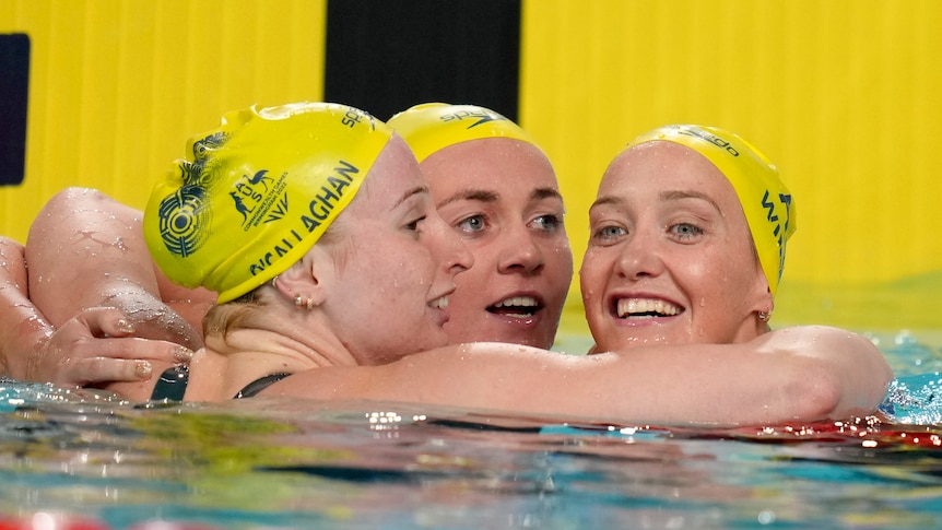 three australian swimmers give each other a hug over the lane rope, all are wearing yellow swimming caps