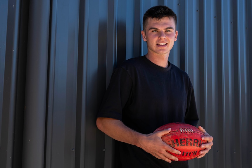 A young football player smiling at the camera with a red AFL football in his hands.