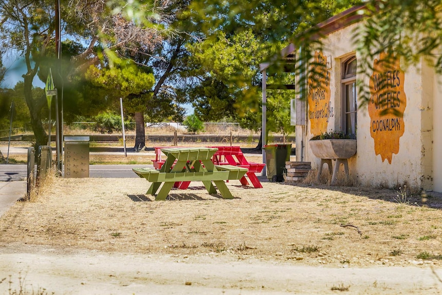 Green and red picnic tables on dry grass next to a stone building with the words 'waffles and jaffles Coonalpyn' painted on.
