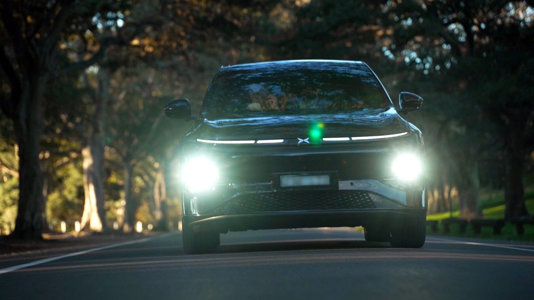 A shiny, modern car drives towards the camera on a tree-lined street. Its headlights are on.