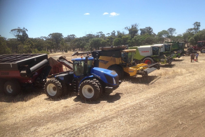 A line up of tractors and harvest vehicles stands in a paddock.