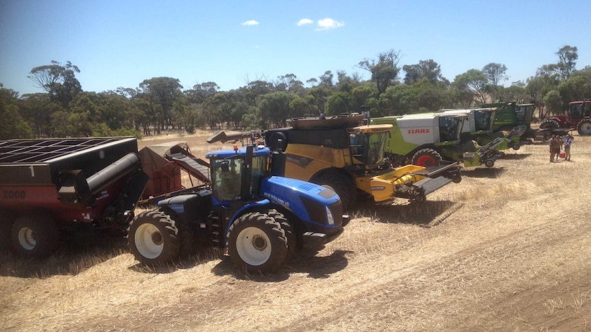 A line up of tractors and harvest vehicles stands in a paddock.