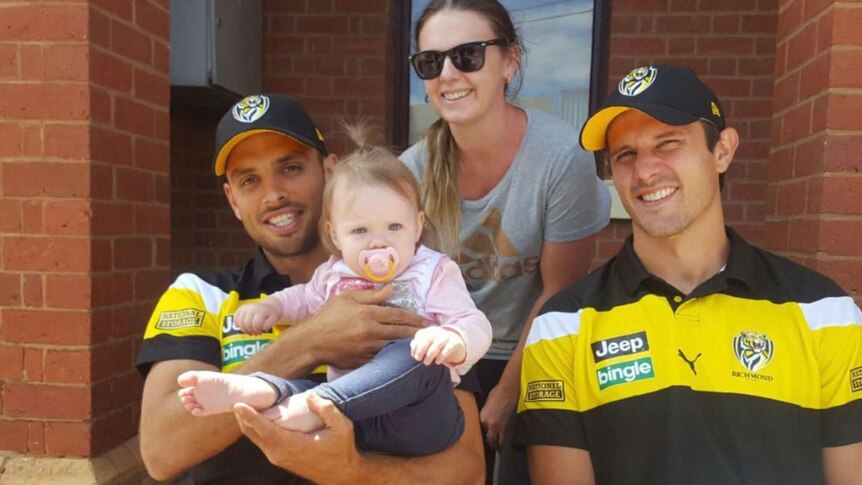 Two Richmond players in caps hold a baby, with the baby's mother sitting behind them.