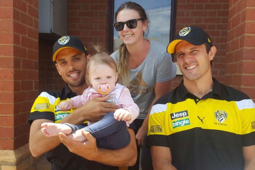 Two Richmond players in caps hold a baby, with the baby's mother sitting behind them.