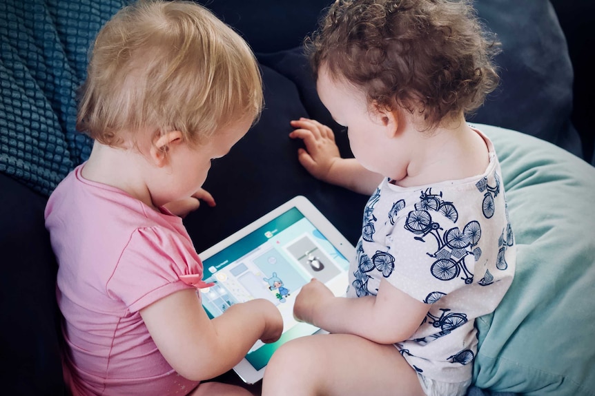 Two toddlers sitting on a couch touch an ipad. On the screen is an app with a cartoon character on it.