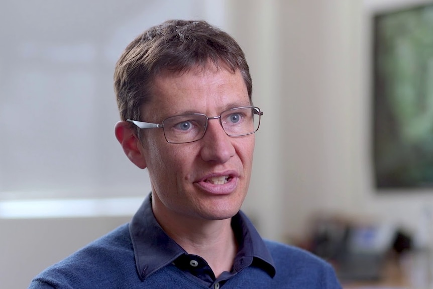 A man being interviewed. He has short, brown hair and glasses. He is wearing a navy blue shirt. Background blurry.