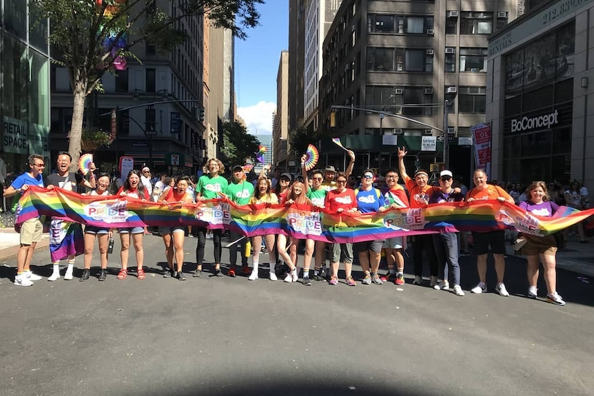 A group of people stand behind a long rainbow colored flag on a New York street. 