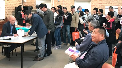 A man sitting at a table signs books as dozens of people line up.