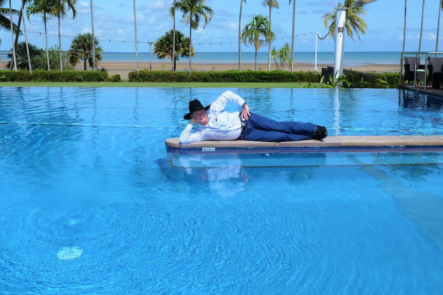 Billy Lowery wearing a cowboy hat, jeans, and shirt lying on a wall in the Mindil Beach Casino pool