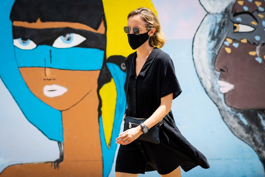 A woman wearing a face mask walks by colourful street art. The woman's dress and mask are both black.