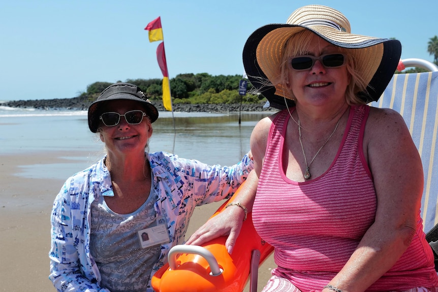 A woman in her 50s in a pink tank top, wearing a sunhat, sitting in a floating wheelchair beside her female carer at the beach.