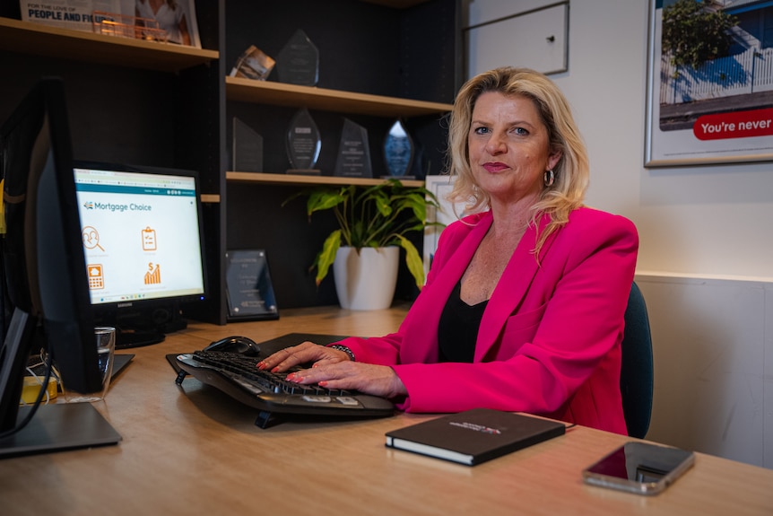 Woman with blonde hair and wearing bright pink jacket sits in front of computer